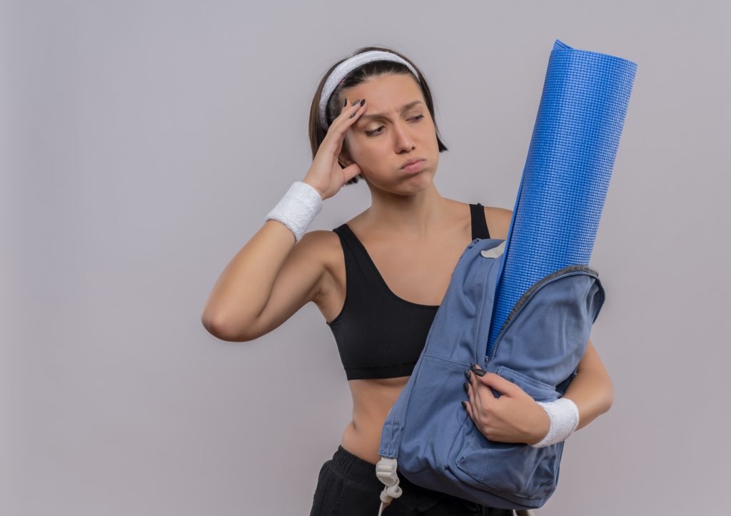 young fitness girl in sportswear hlding backpack with yoga mat looking aside confused and tired blowing cheeks standing over white background